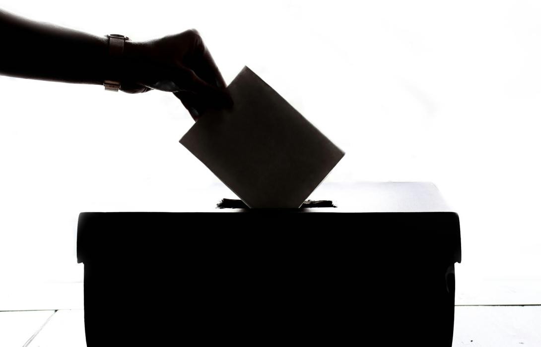 Fotografie einer Hand die einen Umschlag in eine Wahlurne wirft.