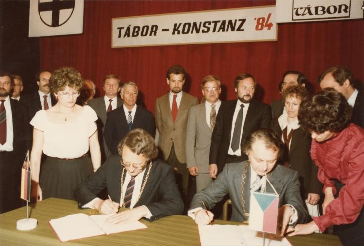 Zwei Männer sitzen an einem Tisch und unterzeichnen jeder ein Schreiben. Um sie herum stehen einige Menschen