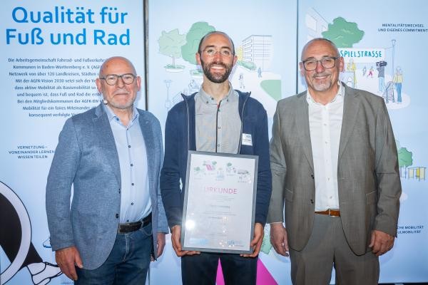 Drei Männer vor einer Infowand; der mittlere hält eine Auszeichnung in den Händen