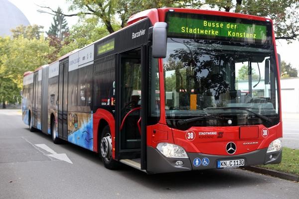 Auf dem Bild ist eine Straße zu sehen. Auf dieser Straße steht ein roter Linienbus der Stadt Konstanz und wartet auf Fahrgäste. Auf der rechten Bildseite ist der Bodensee, ein Stück Wiese und Bäume zu sehen.