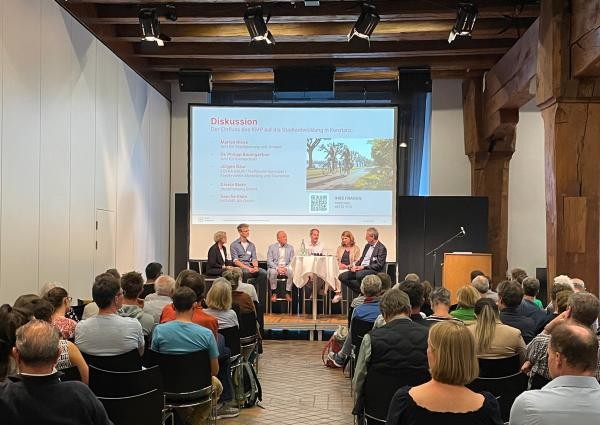 Podiumsdiskussion mit 5 Personen und einem Moderator auf einer Bühne. Im Vordergrund Publikum auf Stühlen
