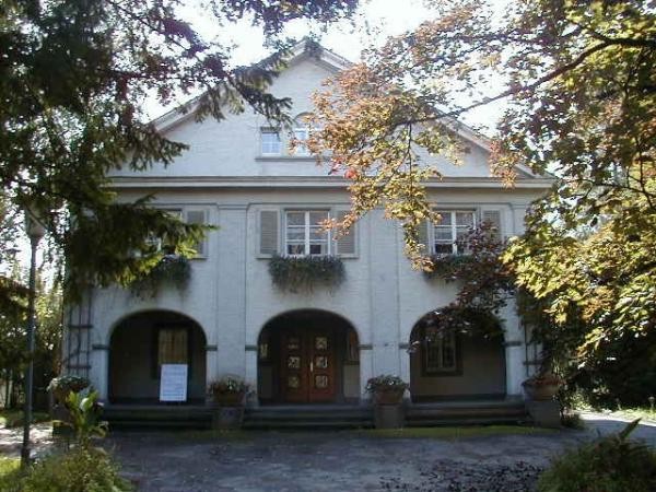 Auf dem Bild zu sehen ist die Fassade eines Hauses, mit drei Rundbögen. Links und rechts wird das Haus im Bild von begrünten Zweigen gerahmt.