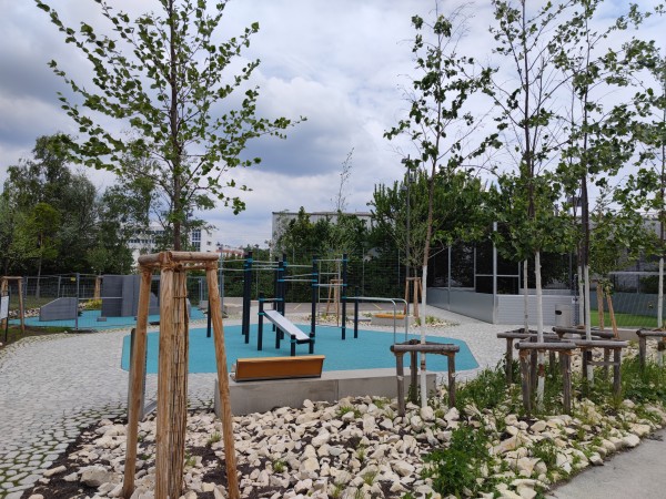 Das Bild zeigt eine moderne Spiel- und Bewegungsfläche in der Grünspange Weiherhof in Konstanz. Zu sehen sind Calisthenics-Geräte auf einem blauen Fallschutzboden, junge Bäume mit Holzstützen, Natursteinflächen und ein Streetballplatz im Hintergrund. Die Anlage ist von Grün umgeben.