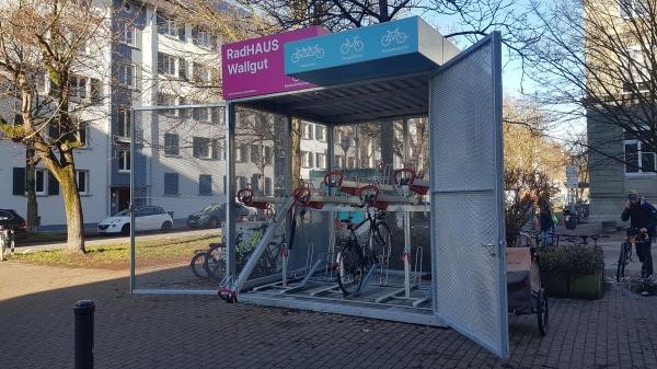 Das Bild zeigt einen überdachten Fahrradabstellplatz namens „RadHAUS Wallgut“ in Konstanz. Die Anlage bietet doppelte Fahrradständer mit oberen und unteren Stellplätzen. Die Türen des Abstellplatzes stehen offen, und ein Schild mit Piktogrammen informiert über die Nutzung.