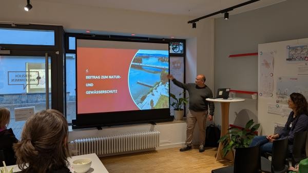 eine Person steht vor einer Personengruppe und hält einen Vortrag mit Präsentation