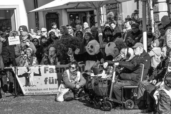 Am Rande eines Fasnachtsumzugs stehen Menschen in Reihen hintereinander. In erster Reihe ist ein Tisch aufgebaut. Daran sitzen drei Menschen mit Mikrofonen. Über ihnen hängt ein Schild mit der Aufschrift Blindenreportage.