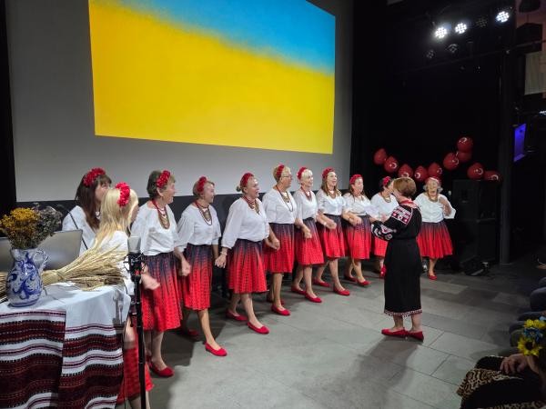 Eine Gruppe von Frauen in traditioneller Kleidung singt auf einer Bühne vor einem Hintergrund in den Farben der ukrainischen Flagge. Die Sängerinnen tragen weiße Blusen, rot-schwarze Röcke, rote Schuhe und Blumenkränze im Haar. Vor der Bühne stehen ein Mikrofon und ein Tisch mit traditionellen Dekorationen.