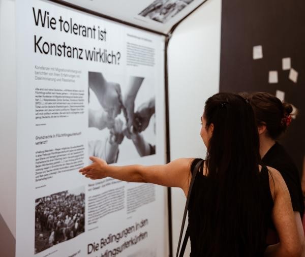Eine Person steht vor einer Wand in der Ausstellung und zeigt auf diese. Man sieht Text und Bilder auf der Wand, sie trägt die Überschrift "Wie tolerant ist Konstanz wirklich?"