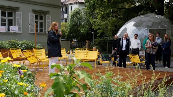 eine Person steht auf einem Platz der mit Hochbeeten gestaltet ist. Sie spricht in ein Mikrophon. Es stehen Menschen darum herum und hören zu.