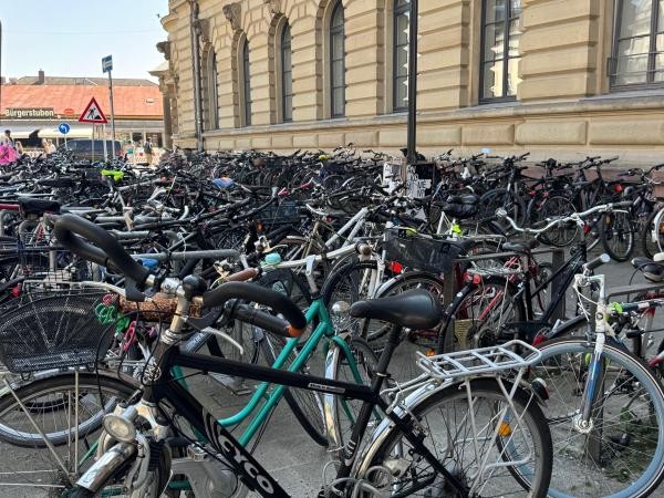 Das Bild zeigt eine große Anzahl eng abgestellter Fahrräder vor einem historischen Gebäude. Im Hintergrund sind ein Straßenschild, Fußgänger sowie das Gebäude „Bürgerstuben“ zu sehen. Die Fahrräder stehen dicht an dicht, teilweise in mehreren Reihen.