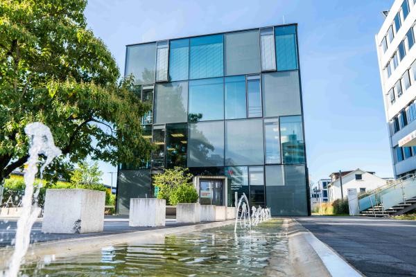 Ein würfelförmiges Gebäude mit Glasverkleidung und einem Brunnen davor