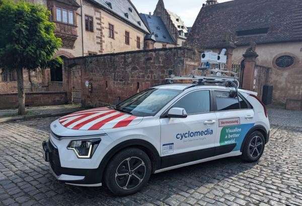 Das Bild zeigt ein weißes Auto mit rotem Warnmuster auf der Motorhaube, das mit Kameras und Sensoren auf dem Dach ausgestattet ist. Auf der Fahrzeugseite steht „cyclomedia – Geo Data Driven Insights“ sowie „Vermessung“. Das Auto steht auf einem Kopfsteinpflaster vor historischen Gebäuden.