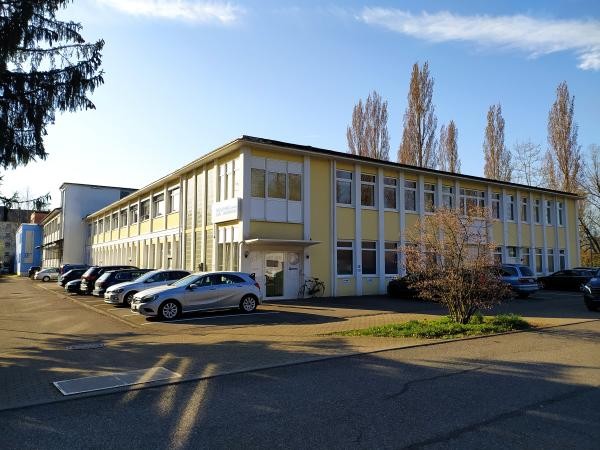 Das Bild zeigt ein zweigeschossiges, gelb-weißes Bürogebäude mit vielen Fenstern. Davor stehen mehrere Autos auf einem Parkplatz, daneben ist ein Fahrrad abgestellt. Im Vordergrund ist ein kleiner Baum mit kahlen Ästen zu sehen.