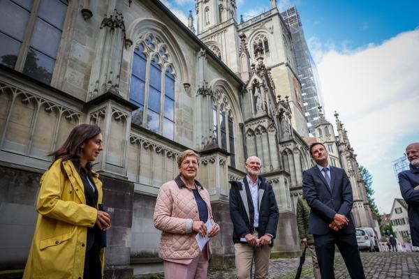Das Bild zeigt eine Gruppe von fünf Personen, die vor einem historischen Kirchengebäude mit neugotischen Fenstern und Verzierungen stehen. Die Personen wirken im Gespräch oder bei einer Führung. Im Hintergrund ist der Turm der Kirche mit Gerüst zu sehen.