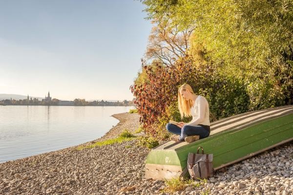 Auf dem Bild ist eine lesende Frau auf einem umgedrehten Ruderboot zu sehen. Das Ruderboot liegt auf Kies neben dem Bodensee. Im Hintergrund erkennt man die Skyline von Konstanz.
