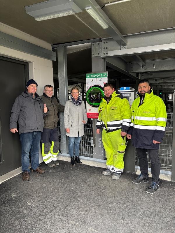Fünf Personen stehen in einem Parkhaus neben einem Defibrillator. Einige tragen neongelbe Sicherheitsjacken.