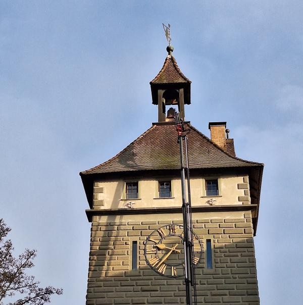 Das Foto zeigt einen Turm aus Stein mit Uhr und kleinem Glockenturm auf dem Dach. Eine Hebebühne reicht bis zur Glocke hinauf. Der Himmel ist blau.