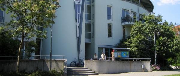 Zu sehen ist ein großes Haus, davor eine Treppe und links und rechts im Bildvordergrund Bäume.