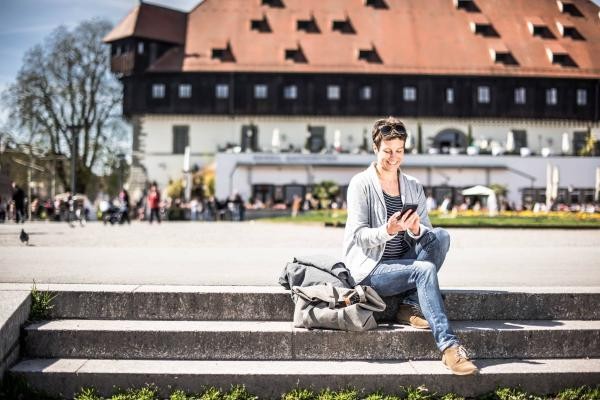 Frau sitzt auf Stufen im Konstanzer Hafen und hält ein Handy in der Handy