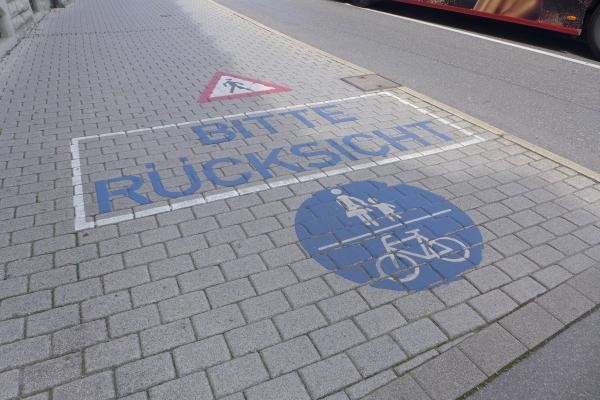 Blaue Bodenmarkierung auf grauem Pflasterstein mit der Aufschrift "BITTE RÜCKSICHT" und Piktogrammen für Fußgänger und Radfahrer in einem blauen Kreis. Im oberen Bereich befindet sich ein Warnschild mit dem Symbol für spielende Kinder. Die Markierung weist auf gemeinsame Nutzung des Verkehrsraums durch verschiedene Verkehrsteilnehmer hin.