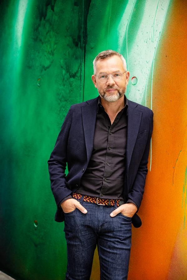 Mann mit Brille, grau-weißen kurzen Haaren und braun-weißem Bart steht vor einer grün-orange eingefärbten Wand. Er hat die Hände in die Taschen seiner Jeans gesteckt und trägt ein dunkelblaues Hemd und ein dunkelblaues Sakko