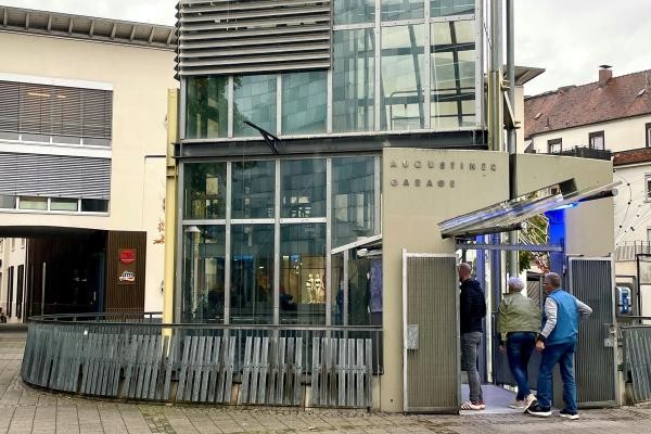 Moderne Gebäudefassade der Augustiner Garage mit Glasfront und gelber Außenwand am Augustinerplatz. Im Vordergrund sind drei Personen zu sehen, die am Eingang mit blauer Beleuchtung stehen, sowie ein holzverkleideter Pflanzkasten. Links im Bild ist ein dm-Drogeriemarkt sichtbar.