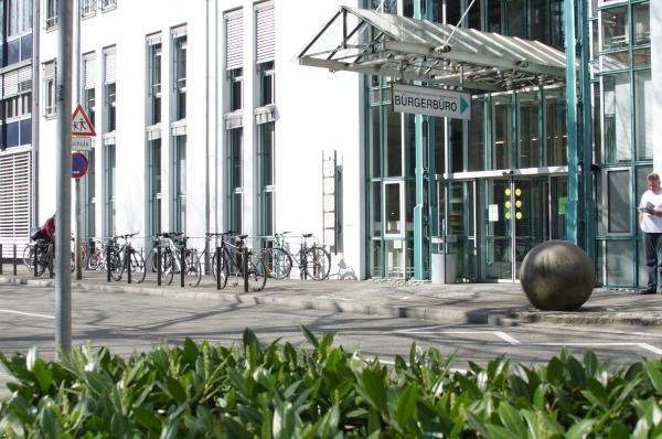 Moderner Glaseingang des Bürgerbüros der Stadt Konstanz mit türkisfarbenen Rahmenelementen und einem transparenten Vordach. Das Gebäude hat eine weiße Fassade mit großen Fenstern, vor dem Eingang liegt eine gepflasterte Fläche mit einer dekorativen Steinkugel. Copyright: Stadt Konstanz.