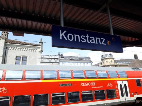 Bahnsteig am Konstanzer Bahnhof mit blauem Stationsschild "Konstanz" und einem rot-weißen Doppelstock-Regionalzug der Schwarzwaldbahn (REGIO DB). Im Hintergrund sind historische Gebäude der Konstanzer Altstadt mit charakteristischen Türmen und Fassaden zu sehen.