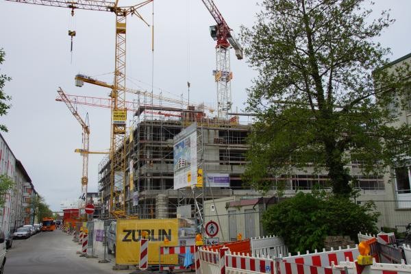 Großbaustelle mit mehreren Baukränen und eingerüstetem Rohbau eines mehrstöckigen Gebäudes. Im Vordergrund sind Bauabsperrungen, Warnschilder und eine gelbe Tafel mit der Aufschrift "ZÜBLIN" zu sehen, rechts steht ein großer Laubbaum. Die Straße ist für Fußgänger gesperrt, mehrere Baufahrzeuge sind im Einsatz.