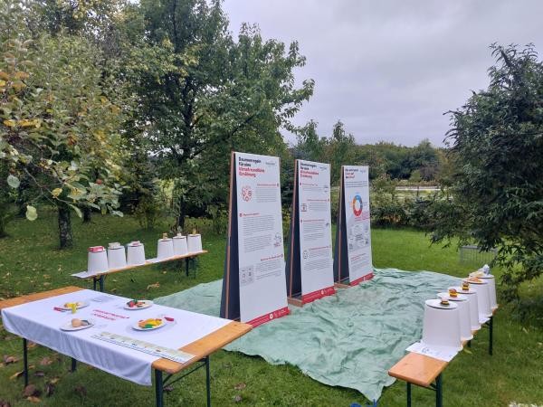 Drei Roll-ups stehen auf einer Folie in einem Garten; von oben hängen Zweige ins Bild. Um die Roll-ups herum steht ein Tisch mit einer Tischdecke und zwei Bierbänke mit weißen Eimern darauf.