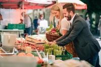 Eine Frau in braunem Rock und weißem T-Shirt und ein Mann in Anzug stehen an einem gut gefüllten Marktstand voll Gemüse und Obst. Sie greifen in eine Kiste Erdbeeren.