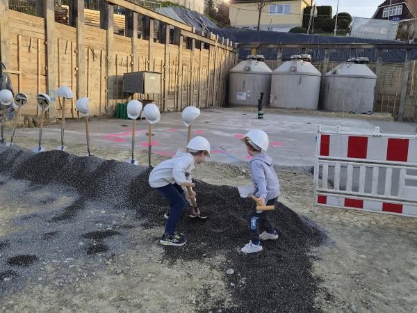 Zwei Kinder mit weißen Bauhelmen führen symbolisch den Spatenstich für die Grundschule Wollmatingen durch und graben mit Holzspaten in einem Erdhügel. Im Hintergrund stehen weitere Bauhelme auf Holzstäben, Holzverschalungen, Baucontainer und eine rot-weiße Bauabsperrung. Die Szene zeigt den Baubeginn des Schulprojekts.