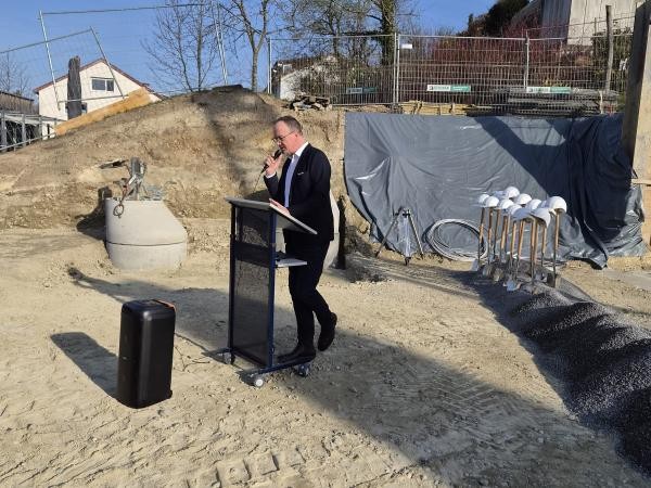Mann in Anzug hält eine Rede am Rednerpult auf einer Baustelle; rechts im Bild liegen Spaten mit weißen Bauhelmen aufgereiht und Materialhaufen für den Spatenstich der Grundschule Wollmatingen.