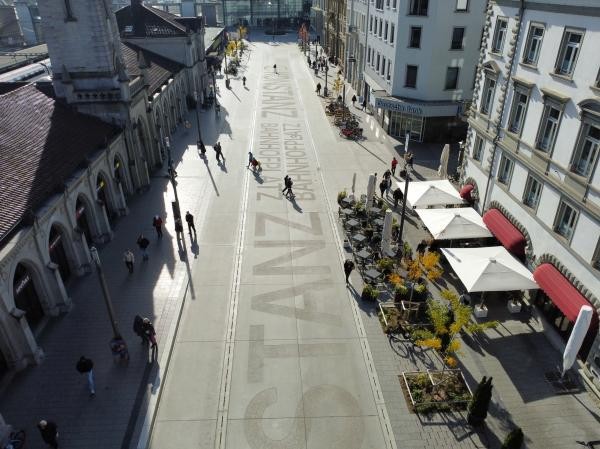 Luftaufnahme des Konstanzer Bahnhofplatzes: Der weitläufige Platz mit hellem Pflasterbelag trägt den Schriftzug „Konstanz Bahnhofplatz" im Boden. Links das historische Bahnhofsgebäude mit Arkaden, rechts Geschäftshäuser und ein Straßencafé mit weißen Sonnenschirmen. Vereinzelte Passantinnen und Passanten überqueren den sonnigen Platz.