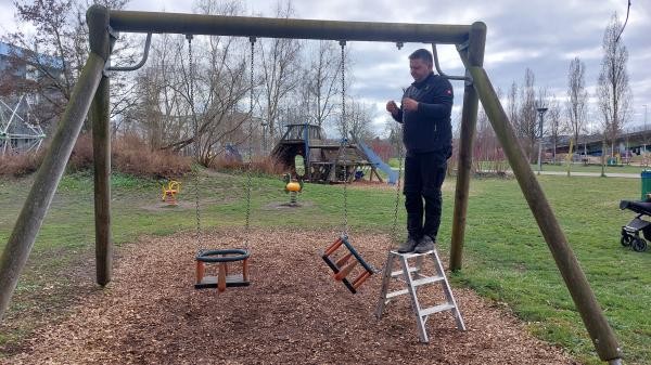 Copyright Stadt Konstanz Ein Mann in einer blauen Fleecejacke steht vor einem großen Holzspielgerät in Form eines Schiffs auf einem Spielplatz. Er hält einen Schraubendreher in der rechten Hand und sticht damit in eine Holzverbindung an der Dachkante des Geräts, um das Holz auf Fäulnis zu prüfen. Im Hintergrund sind kahle Bäume, eine Grünfläche und ein bewölkter Himmel zu sehen. Das Holzgerät weist Graffiti auf.