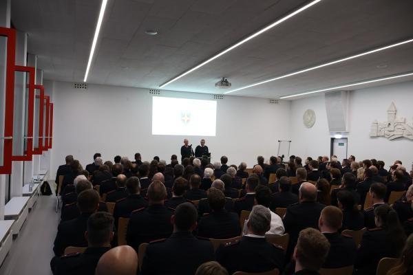 Publikum auf Stühlen in einem Raum; im Hintergrund zwei Redner an einem Stehpult