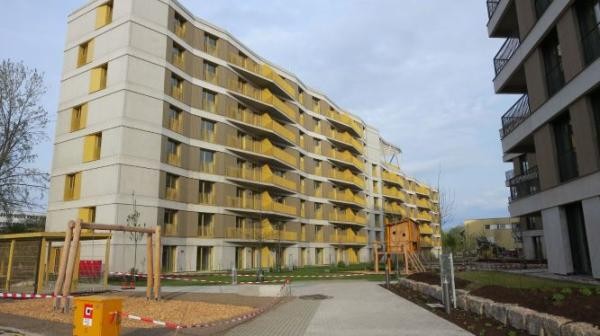 Modernes mehrgeschossiges Wohngebäude im Quartier Weiherhof mit gelben Balkonelementen und geschwungener Fassade. Im Vordergrund ein neu angelegter Innenhof mit Spielgeräten und Grünflächen.