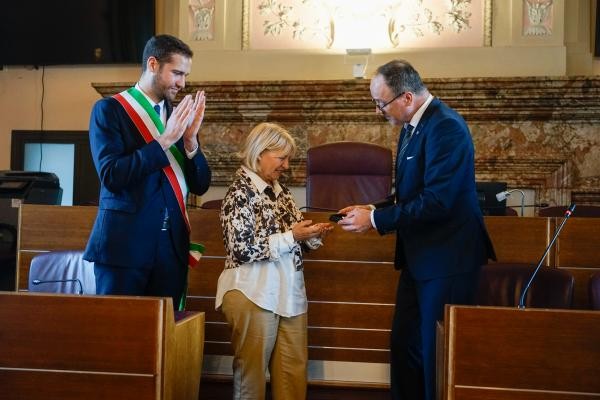 Ein Mann links klatscht und hat eine Scherpe mit der Flagge Italiens darauf, in der Mitte eine Frau mit heller Kleidung, die von einem Mann rechts von ihr etwasüberreicht bekommt.
