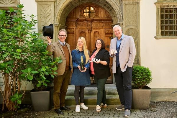 Vier Personen vor einer historischen Holztür, in den Händen halten sie klassische Hochzeitssymbole wle