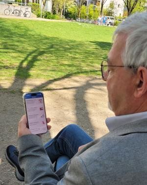 Das Foto zeigt einen Mann mit grauen Haaren, Brille und grauem Jackett. Ersitzt auf einer Bank und hat ein Handy in der Hand - man sieht den schwarzen Umriss des Handys sowie den Bildschirm mit weißen Elementen.