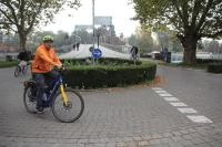Das Foto zeigt bei grauem Wetter einen älteren Mann auf einem Elektro-Fahrrad. Er fährt vor dem Hintergrund einer Brücke um einen kleinen Kreisel, trägt eine grelle orange Jacke und einen Helm, sowie eine Schutzbrille und einen Schnurrbart.