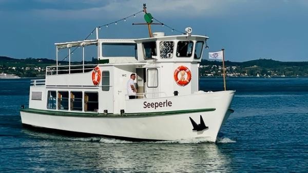 Das Fahrgastschiff „Seeperle" mit weißem Rumpf und Sonnendeck fährt auf dem Bodensee. Ein Besatzungsmitglied steht am Bug, im Hintergrund sind das Ufer und Hügel zu erkennen.