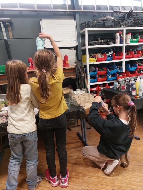 Drei Kinder gestalten in einer Werkstatt bemalte Einmachgläser als Kreativprojekt. Ein Mädchen hält ein blau bemaltes Glas hoch, während im Hintergrund Regale mit Werkzeug und Kleinteilen zu sehen sind.