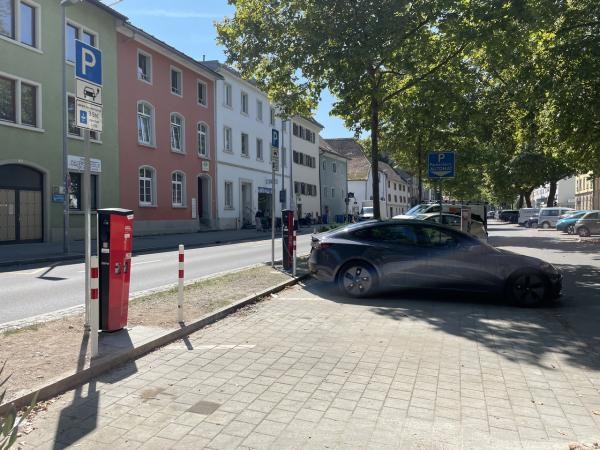 Parkplätze, die mit Stromladesäulen ausgestattet sind. Im Hintergrund eine Häuserzeile, Bäume und parkende Autos.
