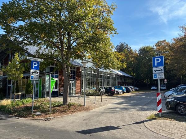 Südparkplatz beim Riesenbergweg 43–49 mit Parkschildern (2 Std., 6–18 Uhr bzw. 8–18 Uhr), im Hintergrund mehrere geparkte Autos vor einem verglasten Gebäude mit Holzverkleidung, umgeben von herbstlich gefärbten Bäumen.