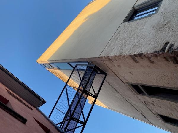Extremer Blick von unten auf den Turm zur Katz in Konstanz: Die abendsonnenbeschienene Fassade des historischen Turms erstreckt sich schräg in den wolkenlosen blauen Himmel, davor eine schwarze Gittertreppe als Notausstieg.