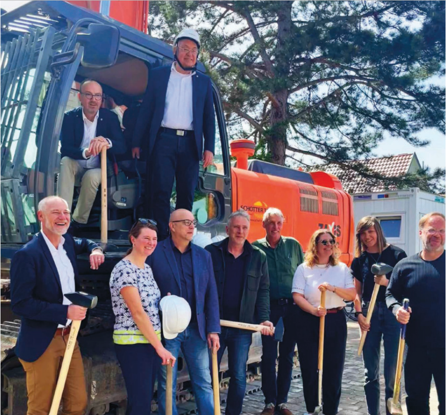 Verschiedene Projektbeteiligte mit Vorschlaghammer in der Hand und einem Bagger im Hintergrund