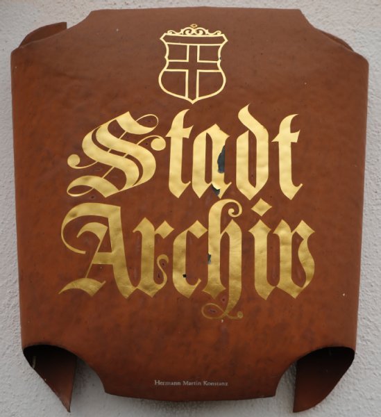 In goldener Farbe steht der Schriftzug Stadtarchiv mit dem Konstanzer Wappen. Das Schild ist rostbraun.