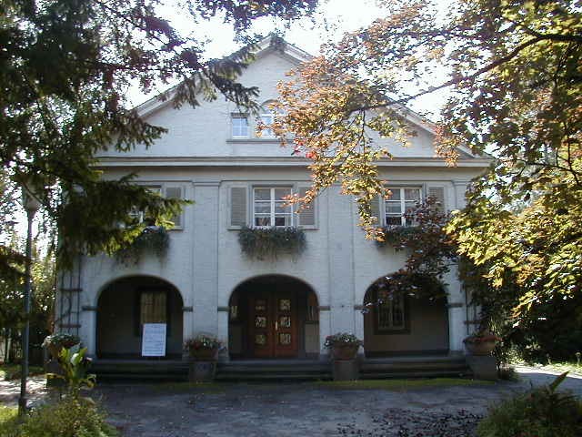 Auf dem Bild zu sehen ist die Fassade eines Hauses, mit drei Rundbögen. Links und rechts wird das Haus im Bild von begrünten Zweigen gerahmt.