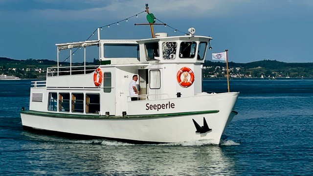 Das Fahrgastschiff „Seeperle" mit weißem Rumpf und Sonnendeck fährt auf dem Bodensee. Ein Besatzungsmitglied steht am Bug, im Hintergrund sind das Ufer und Hügel zu erkennen.
